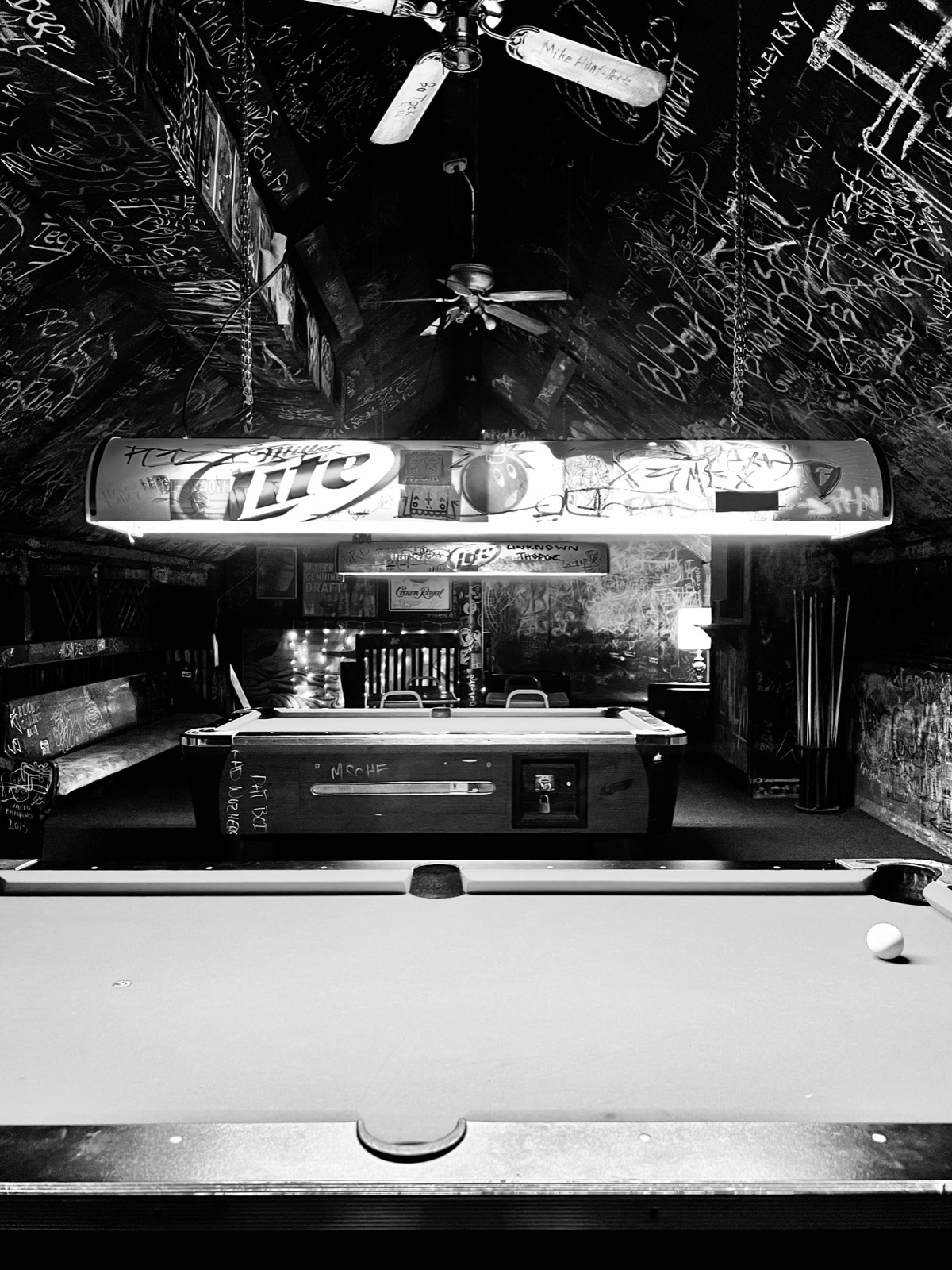 Black and white photo of the back room: pool tables under a hanging beer light, every inch of the ceiling covered in signatures.
