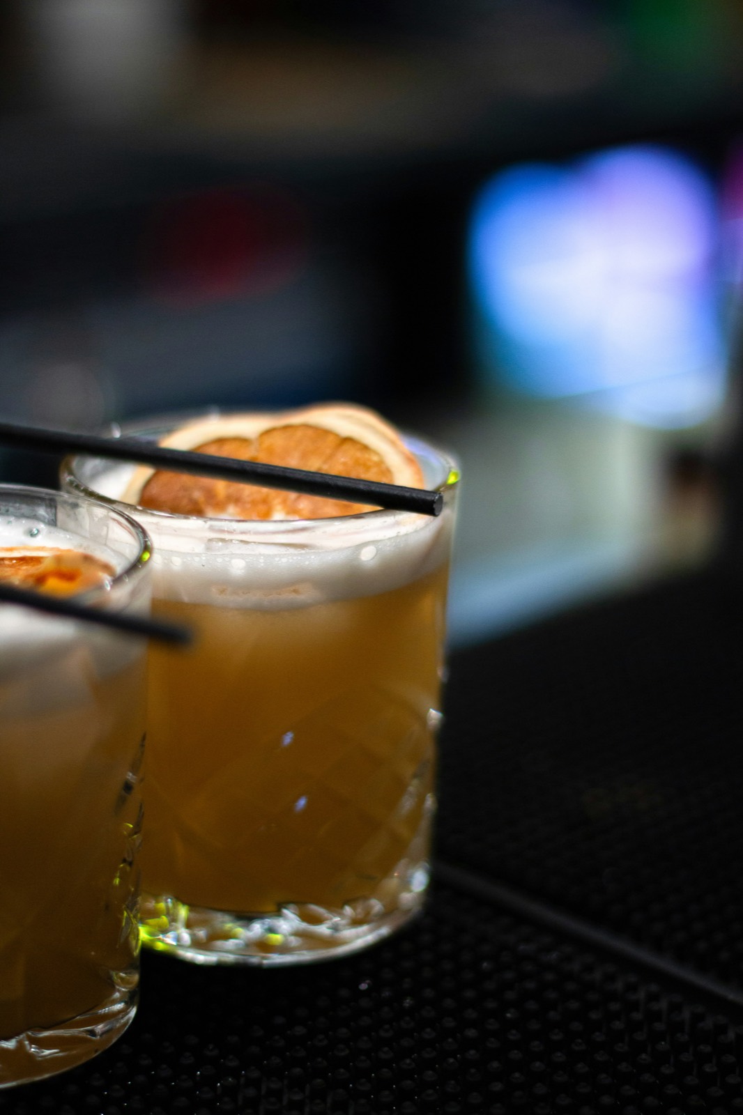 Two cocktails on the bar mat, each with a dried orange wheel and a black straw.