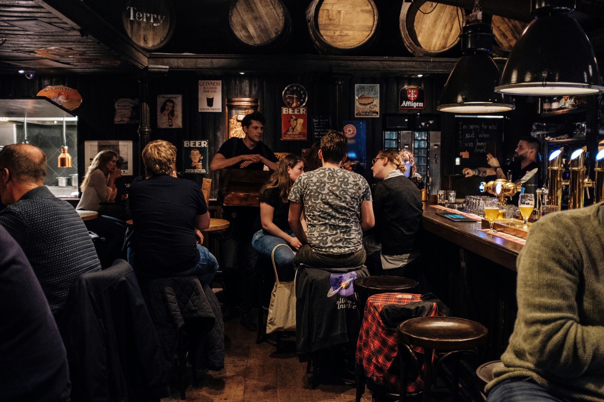 The room on a busy night: regulars on stools along the bar, old beer signs on dark wood walls, barrels overhead.