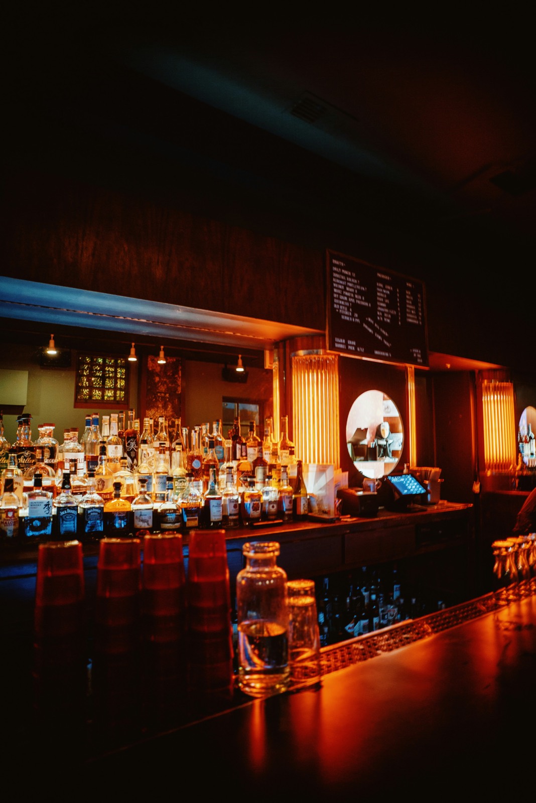 The bar after dark: backlit bottles, a chalkboard menu, and a stack of red cups on the counter.