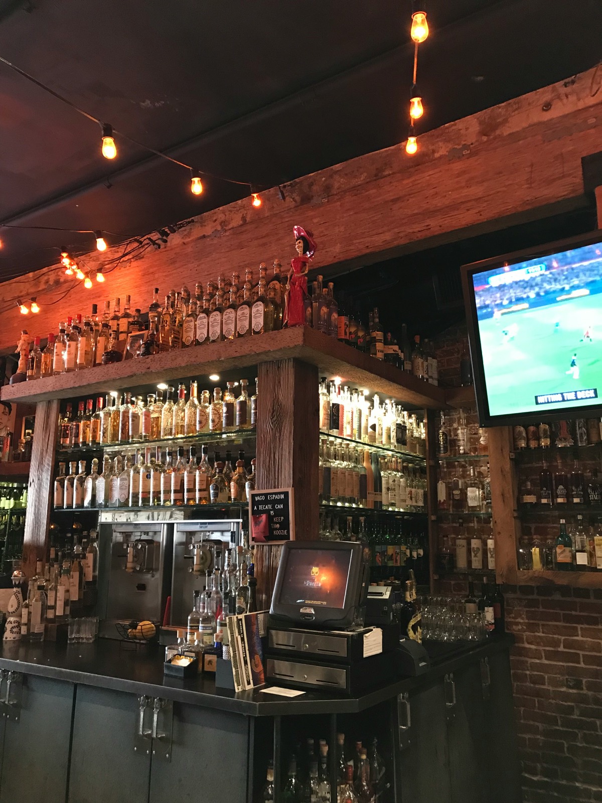 The back bar: shelves of bottles on reclaimed wood, string lights overhead, a game on the TV.